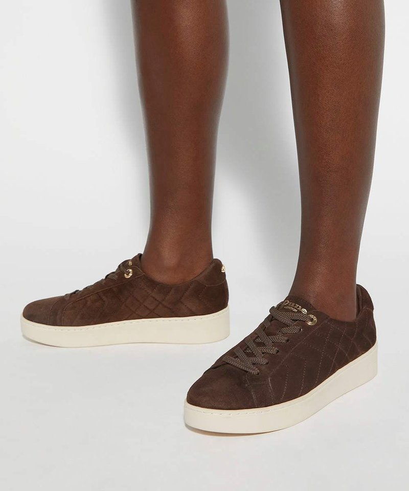 Baskets en daim marron avec motif matelassé et semelles épaisses crème portées sur des jambes à la peau foncée sur fond uni.