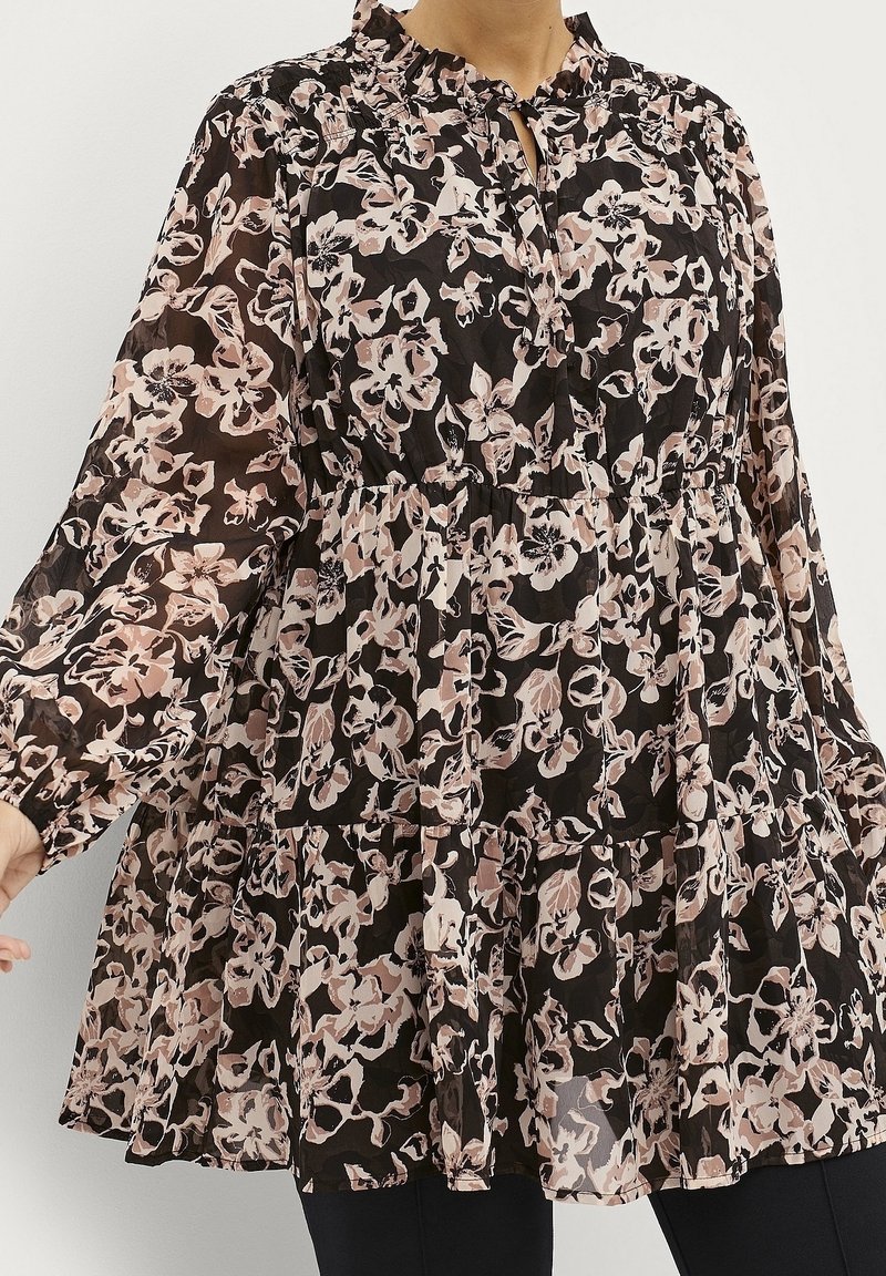 Femme portant un chemisier noir transparent avec des motifs floraux roses et blancs, des manches longues bouffantes et un col à volants.