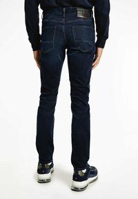 Jeans de mezclilla azul oscuro con un corte slim, costuras visibles y dos bolsillos traseros. Llevados con zapatillas negras y blancas. Diseño limpio sin adornos.