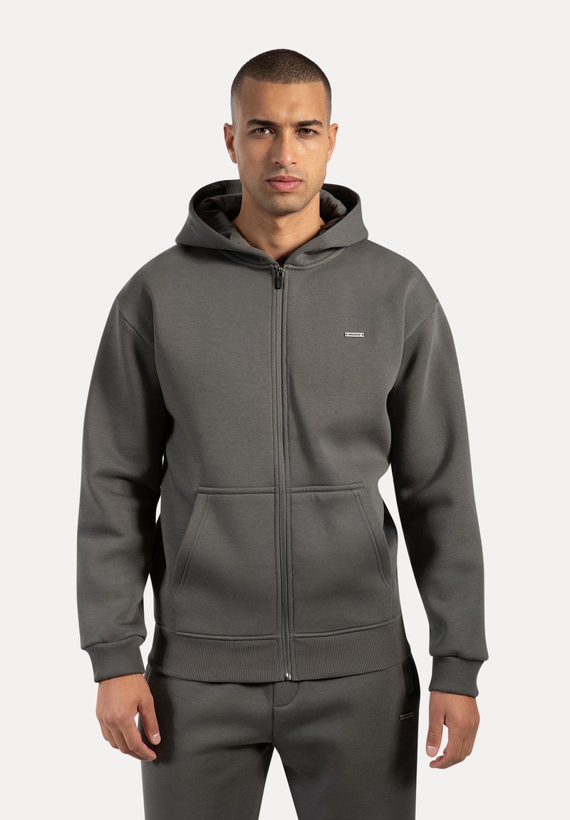 Sudadera con cremallera gris, hecha de tela suave, que cuenta con una gran capucha, bolsillos frontales y puños de canalé. Logotipo pequeño en el pecho.