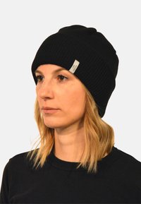 Black knit beanie with a ribbed texture and small woven label. Model wears a black shirt, with shoulder-length blonde hair visible.