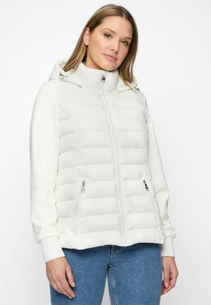 Femme portant une veste matelassée blanche à fermeture éclair avec capuche et poches zippées, associée à un jean bleu, sur un fond uni.