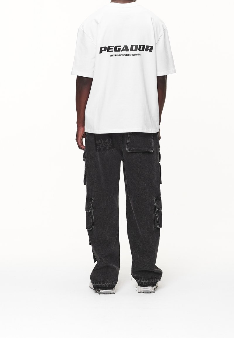 Bílý tričko s logem "PEGADOR" na zádech, spojené s tmavými cargo kalhotami, které mají několik kapes a volný střih.