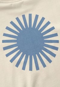 Motif en éclats de soleil bleu avec des rayons régulièrement espacés sur un fond de tissu beige, ressemblant à un soleil stylisé.