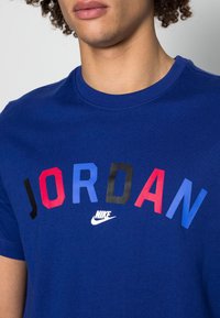 Persona que lleva una camiseta azul con el texto colorido "JORDAN" y un logotipo de Nike blanco centrado debajo del texto.