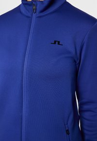 Giacca blu con zip ad alta collo, texture liscia, logo nero sul petto e tasca laterale con zip per funzionalità.
