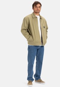 Khaki kleurige jas met een kraag, voorzakken en een patchlogo. Draag het over een wit shirt met een blauwe spijkerbroek en bruine sneakers.