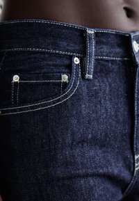 Jeans in denim blu scuro mostrano una vista ravvicinata della vita, con cuciture visibili e dettagli delle tasche arricchiti da finiture in metallo argentato.