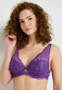 Reggiseno viola in pizzo con motivo floreale, design a scollo a V, bordi smerlati e un piccolo dettaglio metallico al centro. Tessuto morbido e testurizzato.