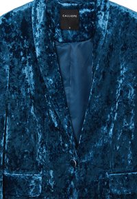 Blazer in velluto teal con collo a punte, chiusura a singolo bottone e due tasche frontali. Fodera interna liscia in una tonalità più scura.