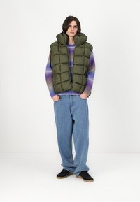 Vesta căptușită verde, cu guler înalt, având secțiuni matlasate. Pur și simplu peste un pulover multicolor. Asortată cu blugi largi de culoare albastră și pantofi negri.