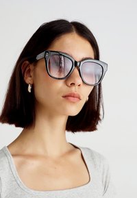 Óculos de sol grandes e pretos com lentes degradê, formato quadrado, detalhes em metal e armações grossas. O modelo usa uma blusa cinza e brincos.