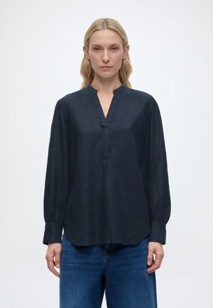 Kobieta stojąca przodem, ubrana w ciemnogranatową bluzkę z długim rękawem i rozcięciem przy dekolcie oraz niebieskie dżinsy, na jednolitym białym tle.