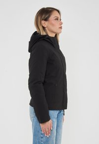 Giacca nera isolata con cappuccio, dotata di zip frontale, tessuto tattili e tasche laterali con zip. Indossata su jeans in denim blu.