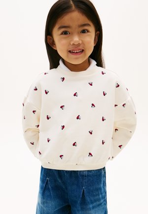 Smilende ung jente med langt mørkt hår som har på seg hvit genser med små røde og svarte hjerte-mønstre og blå dongeribukser.