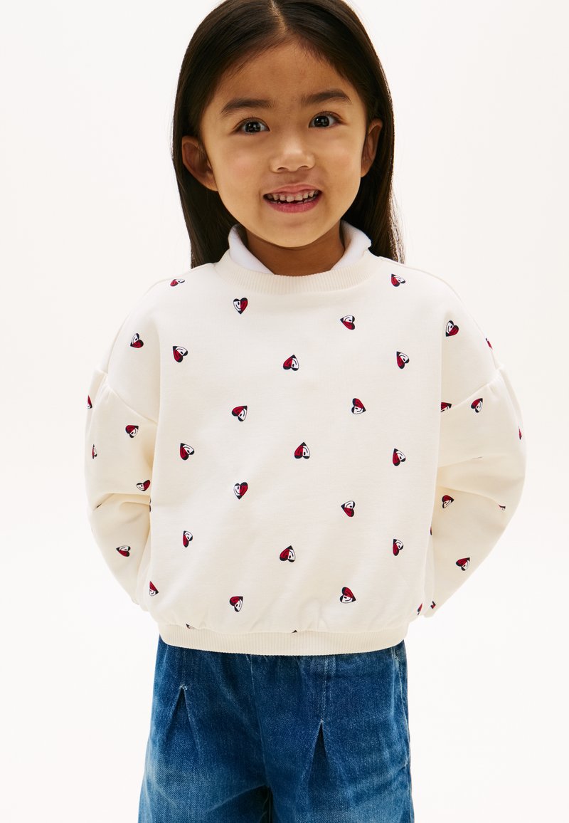 Χαμογελαστή νεαρή κοπέλα με μακριά σκούρα μαλλιά που φοράει λευκό πουλόβερ με μικρά κόκκινα και μαύρα μοτίβα καρδιάς και μπλε τζιν παντελόνι.