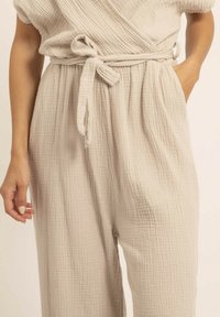 Combinaison en coton beige à enrouler avec une finition texturée et froissée, dotée d'une ceinture à nouer à la taille et d'un design à jambes larges.