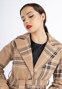 Giovane donna con capelli scuri legati in una coda bassa, indossa un audace rossetto rosso e un cappotto a quadri beige con cintura sopra un top nero, guardando in basso.