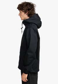 Chaqueta negra con capucha y cordón, bolsillo frontal y puños elásticos. Tela suave y corte relajado, mostrada de lado.