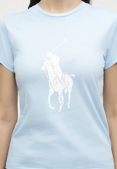 Άτομο που φοράει γαλάζιο πουκάμισο με λευκό γραφικό του παίκτη πόλο σε άλογο που κουνάει ρόπαλο, στο κέντρο του στήθους.