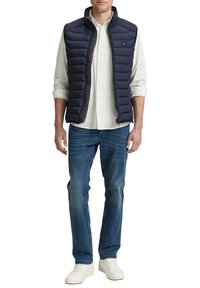 Doudoune sans manches bleu marine à matelassage horizontal, fermeture éclair, et patch logo, portée sur une chemise blanche à boutons et un jean bleu avec des baskets blanches.