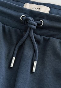 Pantaloni sportivi blu navy con coulisse annodata con terminali metallici e un'etichetta della vita bianca parzialmente visibile.