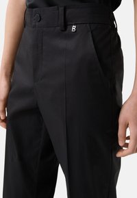 Zwarte broek van gladde stof, met een nette pasvorm, zijzakken, een zichtbare knop en een zilveren accent met de letter "B" op de tailleband.