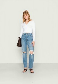 Top branco de mangas longas envolvente, jeans azuis de cintura alta desbotados com rasgões nos joelhos, mala preta de ombro e chinelos pretos. Conjunto simples e casual.