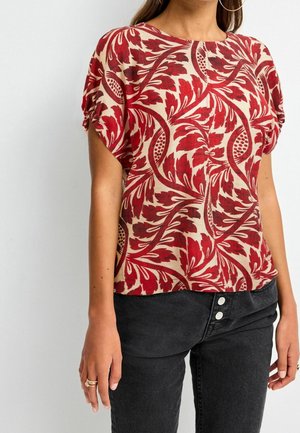 Femme portant un haut à manches courtes rouge et beige à motif floral, un jean noir taille haute et des boucles d'oreilles argentées en anneau.