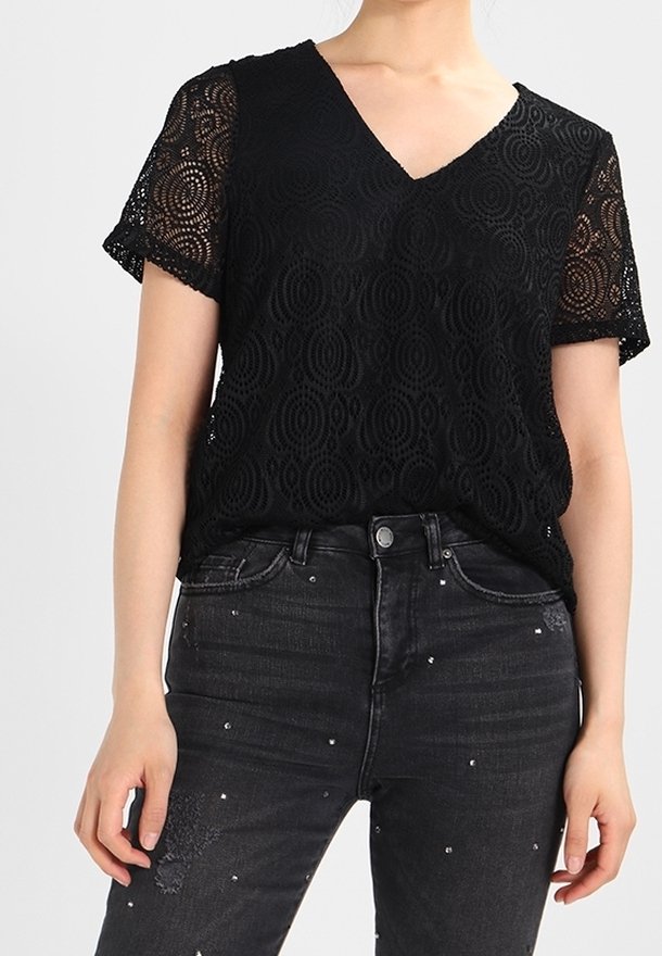 Femme portant un haut noir en dentelle à manches courtes et col en V, et un jean noir taille haute avec de petits points brodés.
