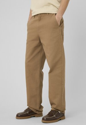 Homme portant un pantalon beige droit et des mocassins en cuir marron, debout les mains dans les poches devant un fond gris.