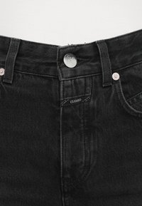 A fekete farmerek klasszikus öt zsebbel rendelkeznek, ezüst gombos záródással és elől egy "CLOSED" címkével. A textúrázott anyag jól látható.