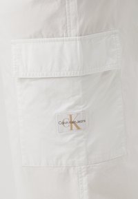 Witte stof met een gladde textuur heeft een opgestikte zak. Een label toont "Calvin Klein Jeans" met een gestileerde "CK" in goud.