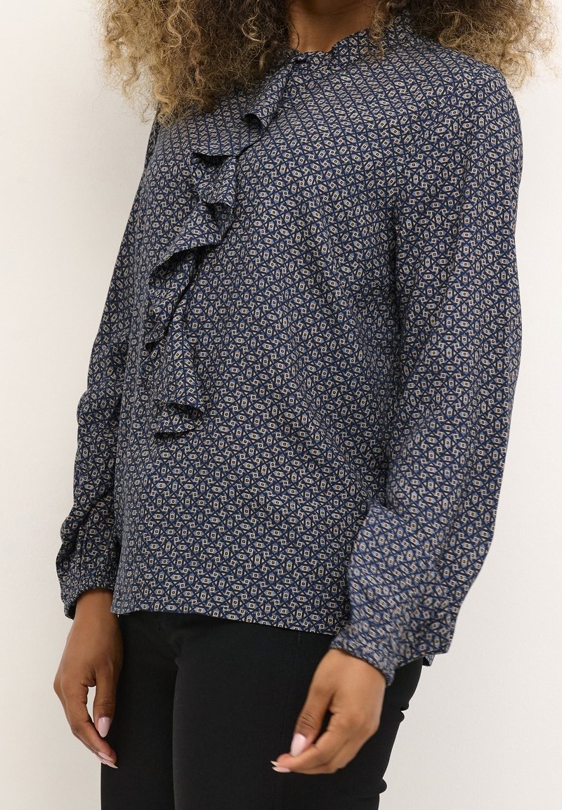 Femme portant un chemisier bleu marine à manches longues avec un motif géométrique beige et un détail volanté, associé à un pantalon noir, debout devant un fond uni.