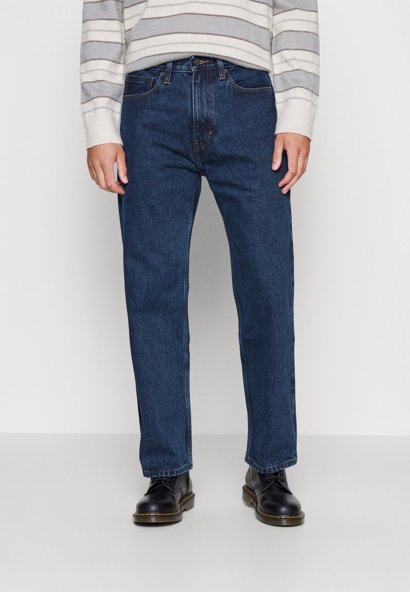 Jeans de mezclilla azul oscuro con corte recto, diseño de cinco bolsillos y detalles de costura sutil, combinados con botas negras de cordones.