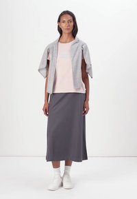 Graue Strickjacke über einem hellpinken Shirt mit Text, kombiniert mit einem langen grauen Rock. Weiße Sneaker, glatte Texturen und lockere Passform.