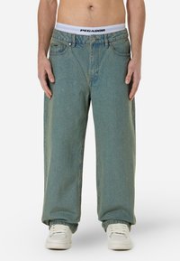 Light blue oversized denim jeans with a high waist, front pockets, and subtle fade. Paired with white sneakers and a branded waistband.