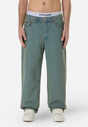 Light blue oversized denim jeans with a high waist, front pockets, and subtle fade. Paired with white sneakers and a branded waistband.