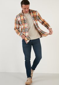 Flanellen overhemd met een geel, rood en grijs geruit patroon, gecombineerd met een beige t-shirt en donkerblauwe skinny jeans. Bruine veterschoenen.