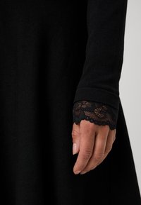 Long-sleeved black dress with a textured fabric. Cuffs feature intricate black lace trim with scalloped edges. Hand is positioned at cuff.