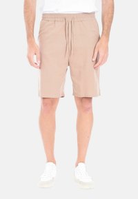 Pantaloni beige chiari con vita elastica e cordoncino, caratterizzati da una texture liscia e una lunghezza fino al ginocchio. Indossati con scarpe da ginnastica bianche.