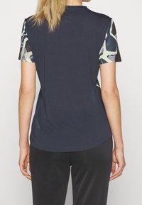 Vrouw met blond haar die een marineblauw shirt met korte mouwen draagt, met groene en zwarte patronen op de mouwen, gezien van achteren.