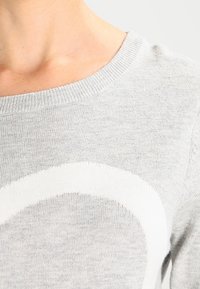 Gros plan d'une personne portant un pull en tricot gris clair avec un motif courbé blanc près de la poitrine et un col rond.