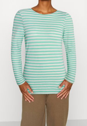 Personne portant une chemise à manches longues rayée horizontalement en vert clair et blanc, avec un pantalon marron, se tenant devant un fond blanc uni.