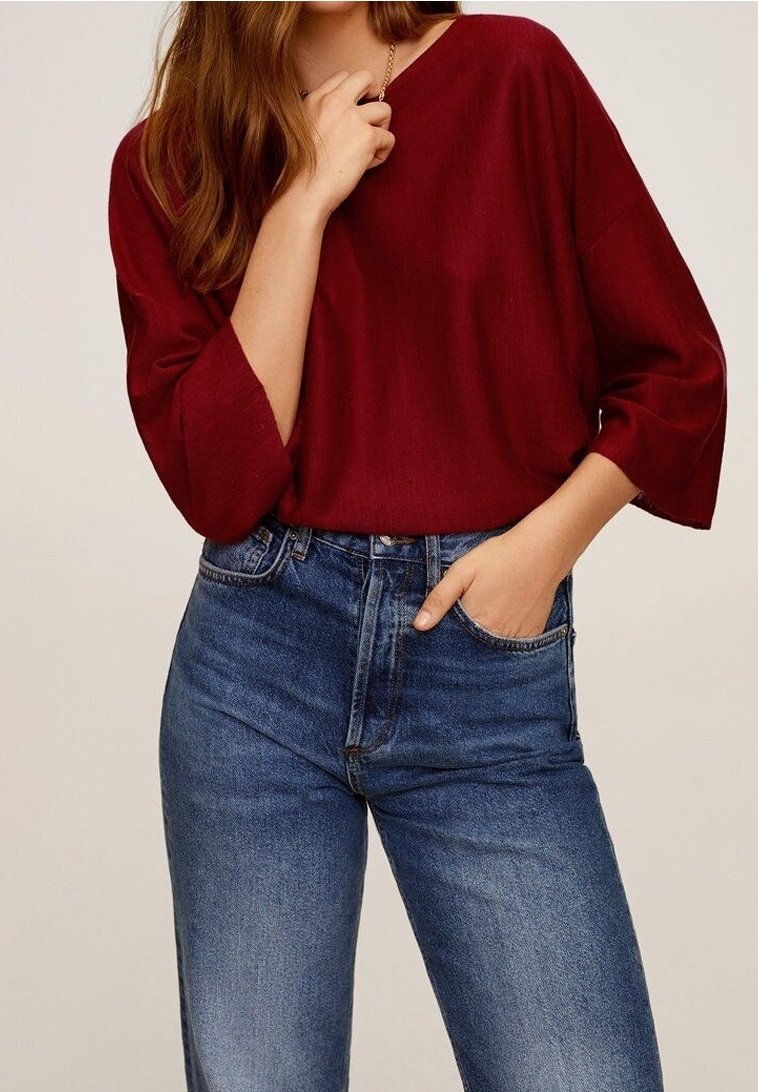 Femme portant un haut rouge ample et un jean bleu taille haute, une main dans la poche et l'autre touchant un collier.