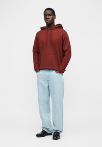 Mężczyzna stojący z rękami w kieszeniach, ubrany w rdzawobrązową bluzę z kapturem, jasnoniebieskie dżinsy i czarne buty na jednolitym białym tle.