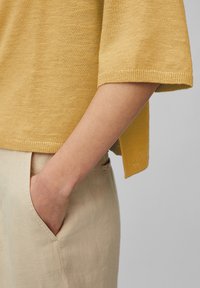 Top en tricot jaune moutarde avec coupe décontractée et manches courtes, associé à un pantalon en lin beige avec une poche sur le côté. Design simple et épuré.