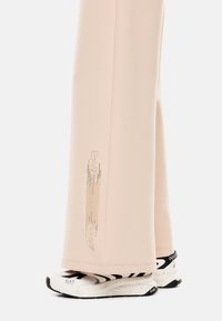 Pantalones de chándal rosa claro con un detalle de logotipo texturizado en el costado, combinados con zapatillas deportivas en blanco y negro con una suela moteada.