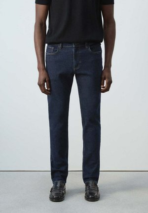 Mann trägt dunkelblaue Slim-Fit-Jeans, schwarzes Hemd und schwarze Lederschuhe und steht vor hellem Hintergrund.