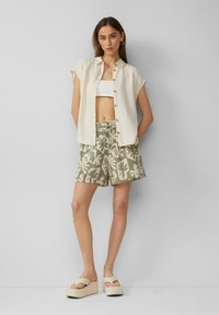 Leichte kremfarbene Knopfhemd über einem weißen Crop-Top, kombiniert mit gemusterten olivgrünen Shorts und Plateausandalen.